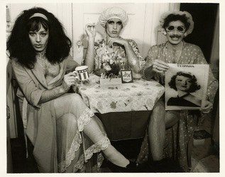Photograph called Tea Time, Three Revolutionaries (Tedde, Jesse, Bobo) - San Francisco, 1974 shows three drag queens seated at a kitchen table posing with an S&H green stamps sign prominently displayed on the table.